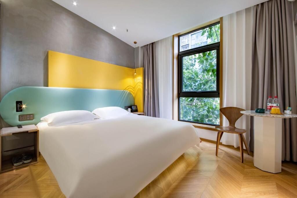 A modern hotel room with a large bed, a desk with a chair, a window with curtains, and a view of greenery outside.