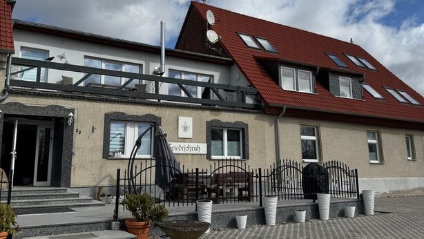Ein Gebäude mit rotem Dach, einem Schild mit der Aufschrift 'Friedrichruh' und einem gepflasterten Innenhof mit Blumentöpfen.