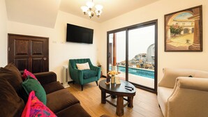 A living room with a brown sofa, a green armchair, a round coffee table, a flat-screen TV, and a large window with a pool view. - Casa de la Abuelita w/Private Pool & Volcano View (San Pedro Las Huertas)