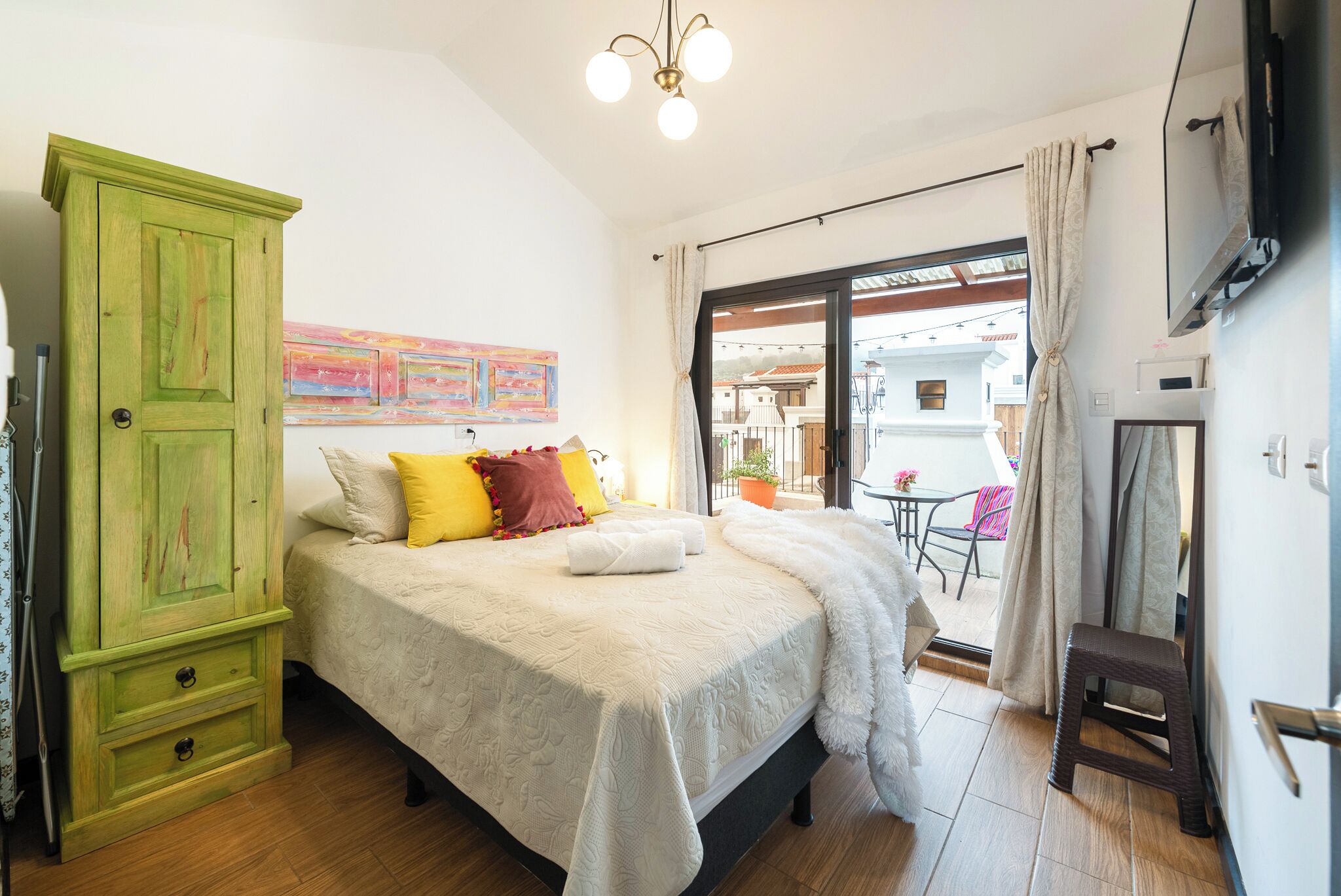 A bedroom with a bed, a green wardrobe, a small stool, and a balcony with a table and chairs.