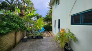A narrow concrete pathway flanked by lush greenery and a white building with windows and a door. - One minute walk to one of the best snorkling/swimming beaches in Rarotonga (Arorangi)