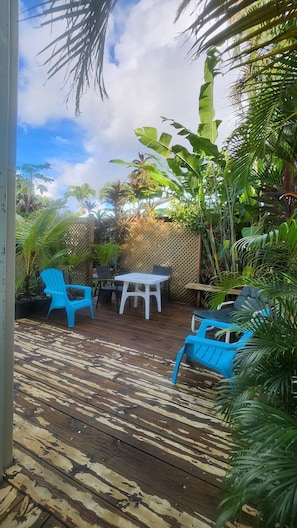 Une terrasse en bois avec une table et des chaises, entourée de plantes tropicales et d’une clôture en treillis de bois.