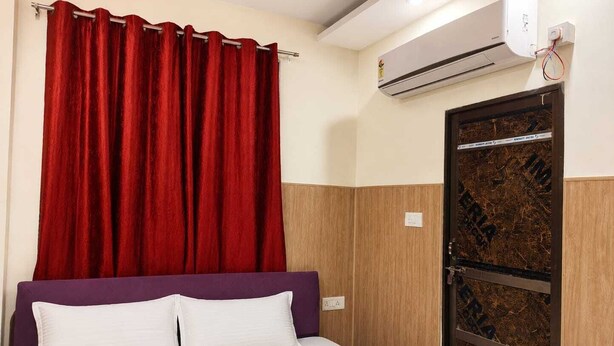 A hotel room with a bed, red curtains, a white pillow, a purple headboard, and an air conditioning unit. - Shree Balaji Paying Guest House (Varanasi)