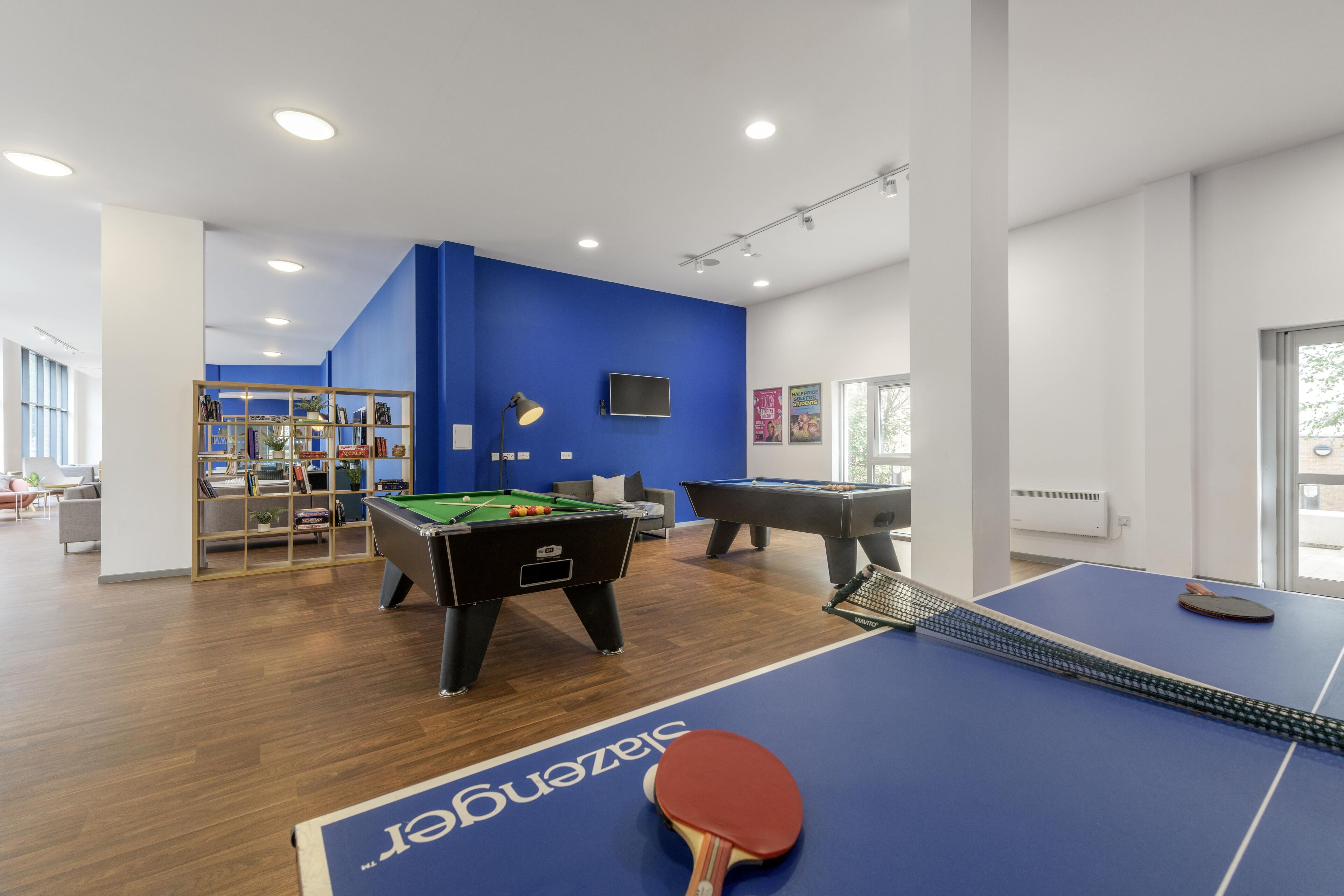 A modern indoor recreational area with pool tables, a ping pong table, and a bookshelf.