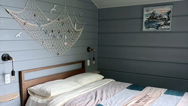 A bedroom with a bed, a hanging net decoration, a framed picture on the wall, and a wooden headboard. - MATRYOSHKA HOUSE (Yoichigun)