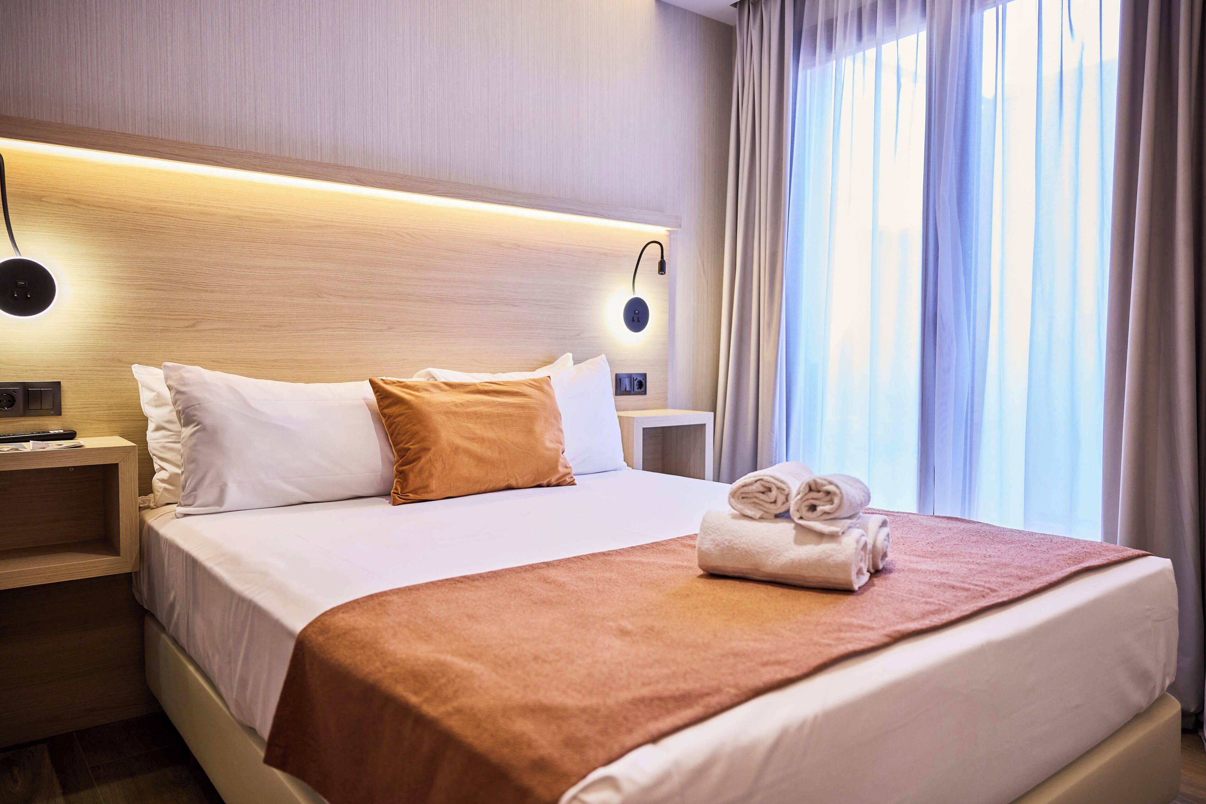 Habitació d'hotel amb un llit, una taula de nit, una làmpada penjada a la paret i una finestra amb cortines.