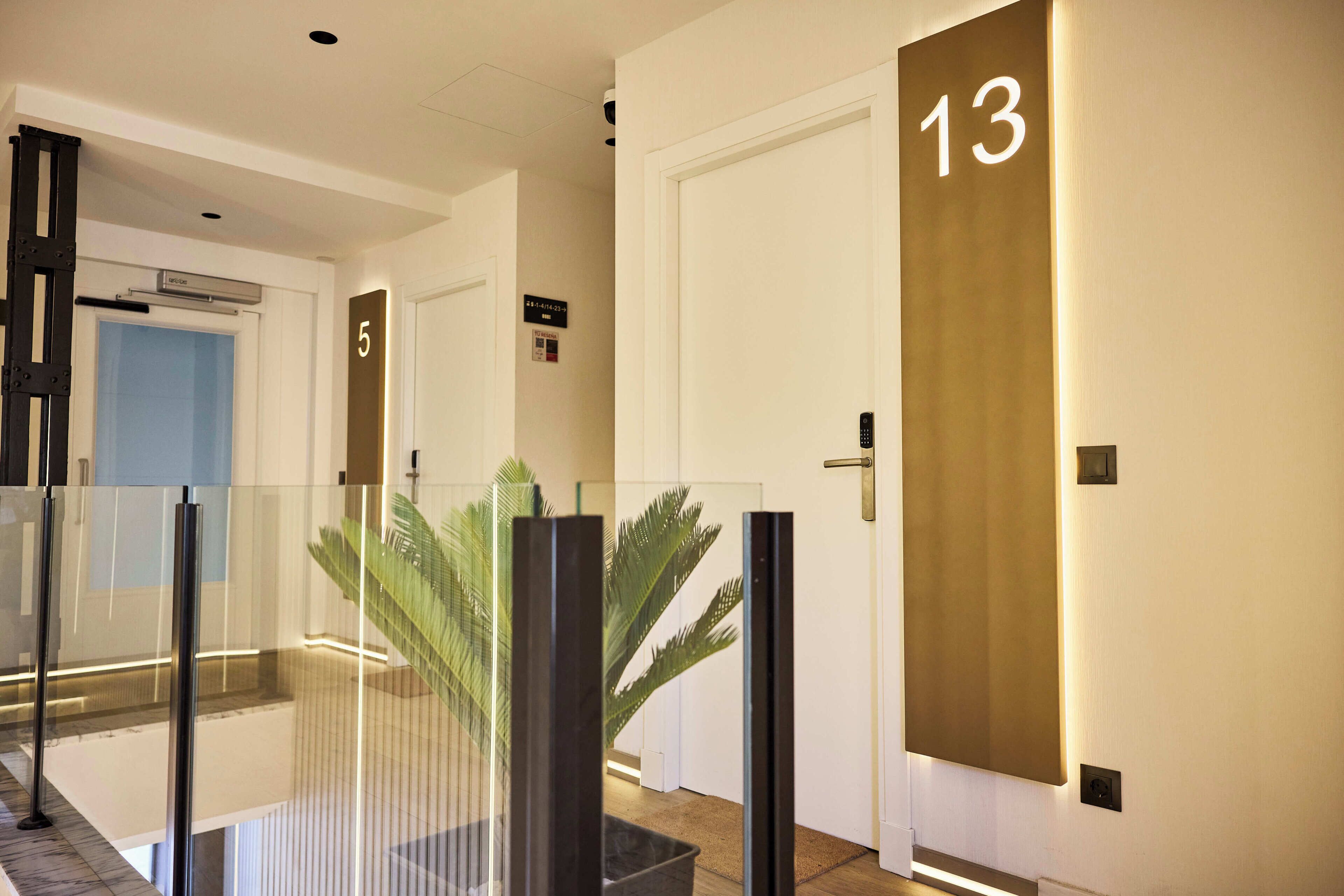 Un pasillo de hotel con barandas de vidrio, una planta de palma y puertas numeradas 5 y 13.