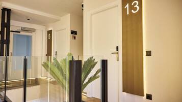Un pasillo de hotel con barandas de vidrio, una planta de palma y puertas numeradas 5 y 13.