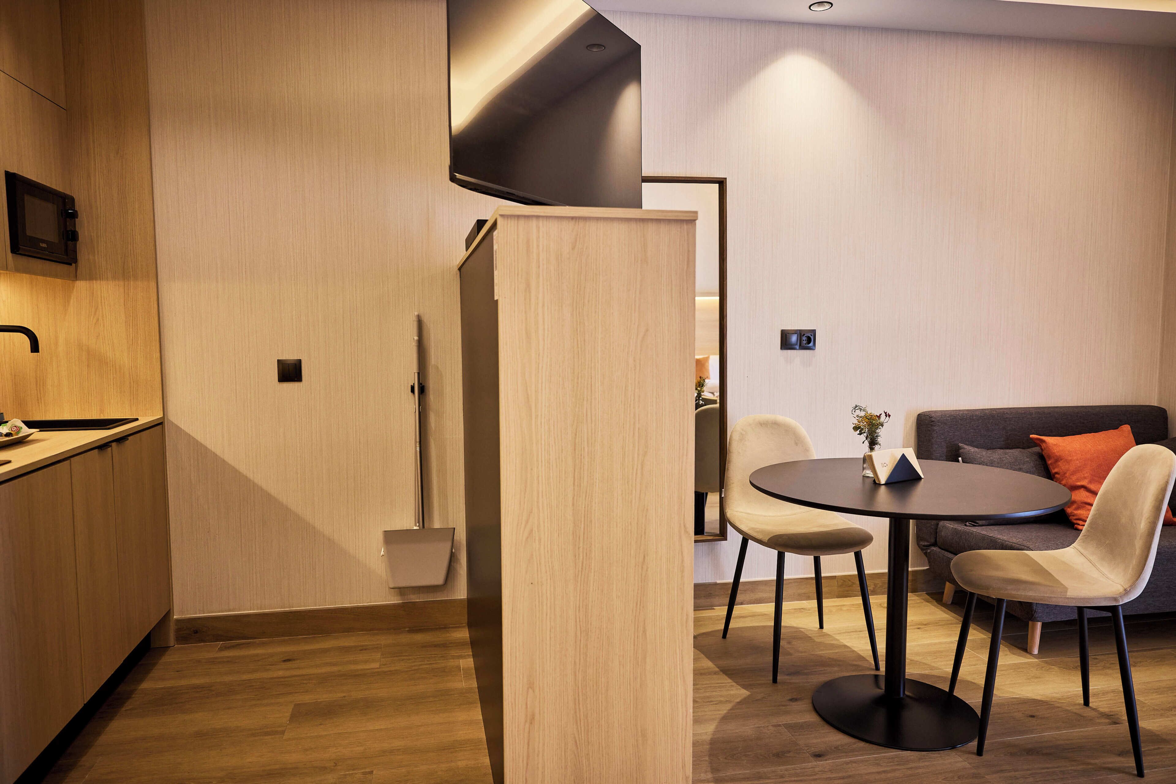 A modern kitchen with wooden cabinetry, a dining area with a round table and chairs, and a sofa in the background.
