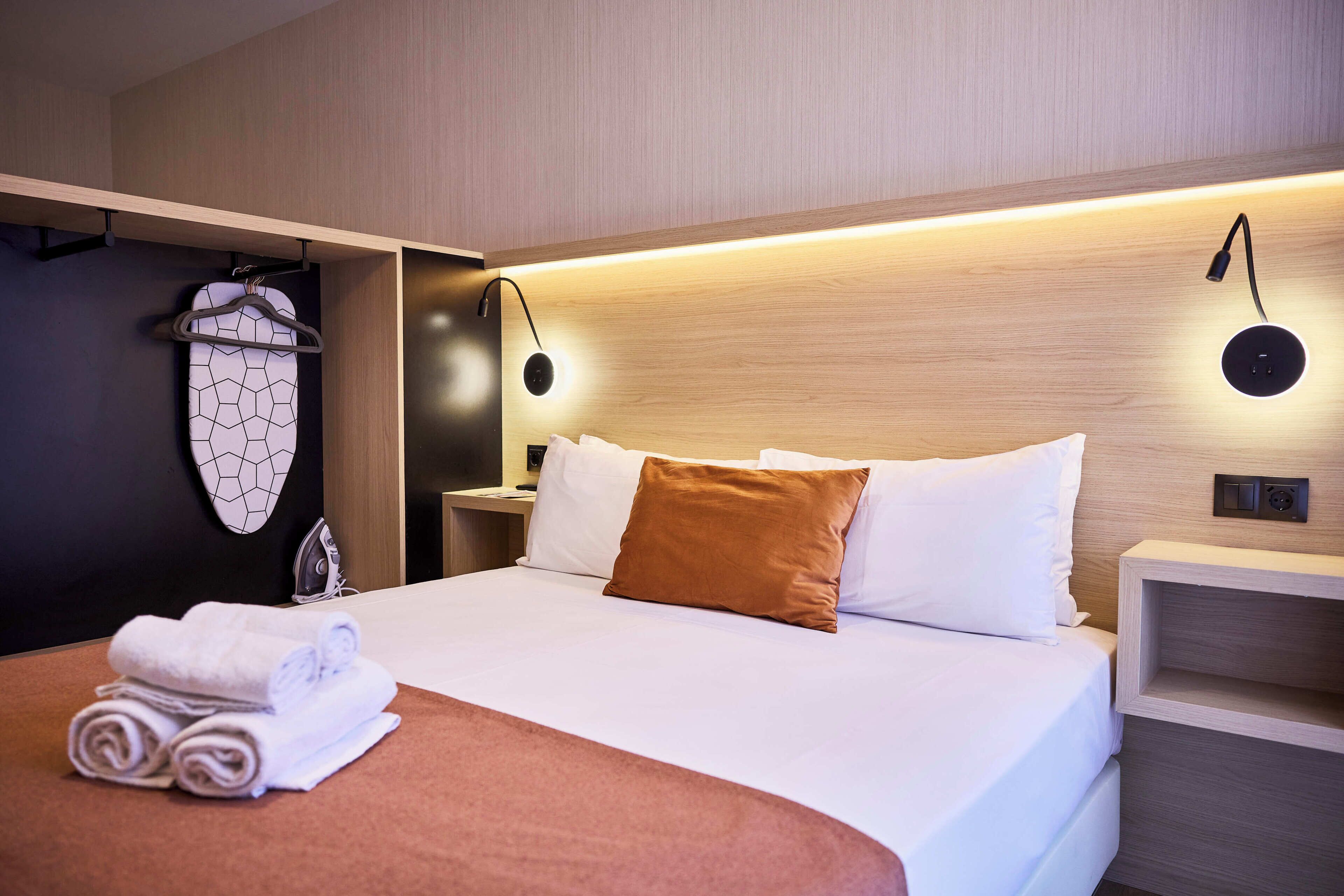 Una habitación de hotel con una cama bien hecha, una percha para ropa, una mesita de noche con lámpara y un enchufe en la pared.