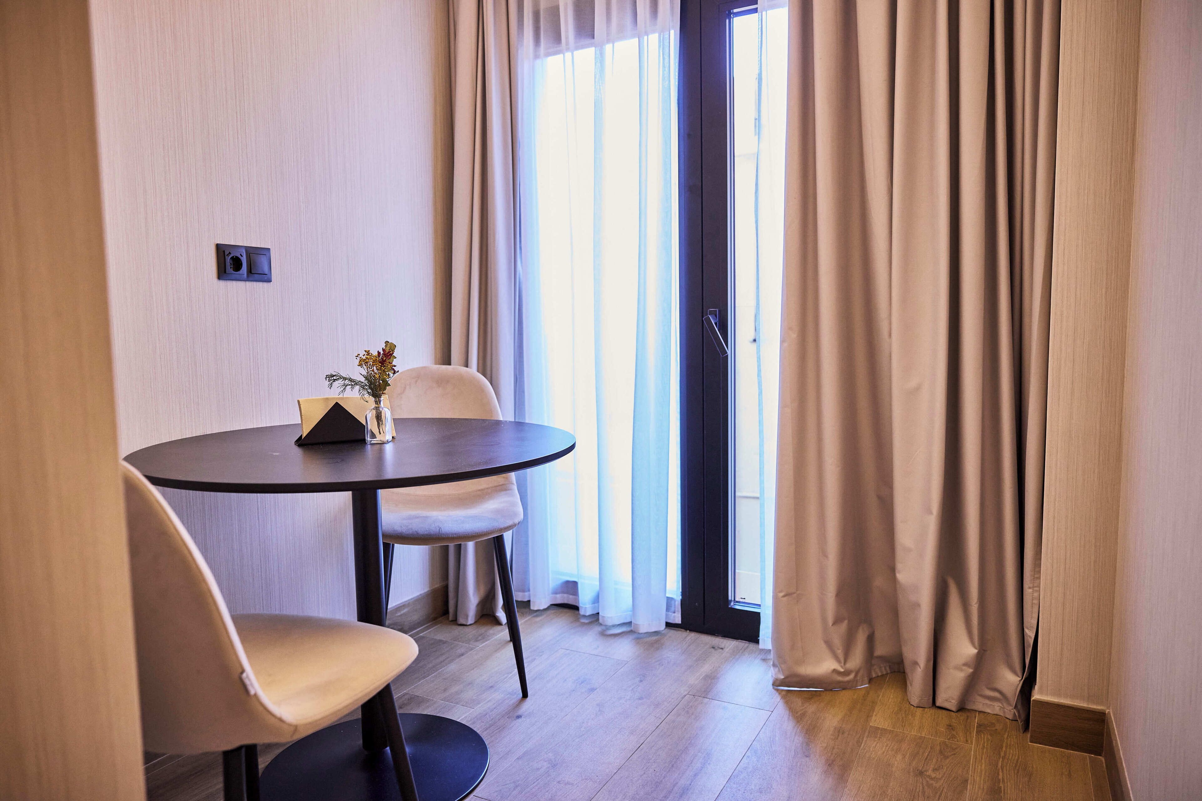 Una habitación con una mesa redonda, dos sillas, un jarrón con flores y una ventana con cortinas.