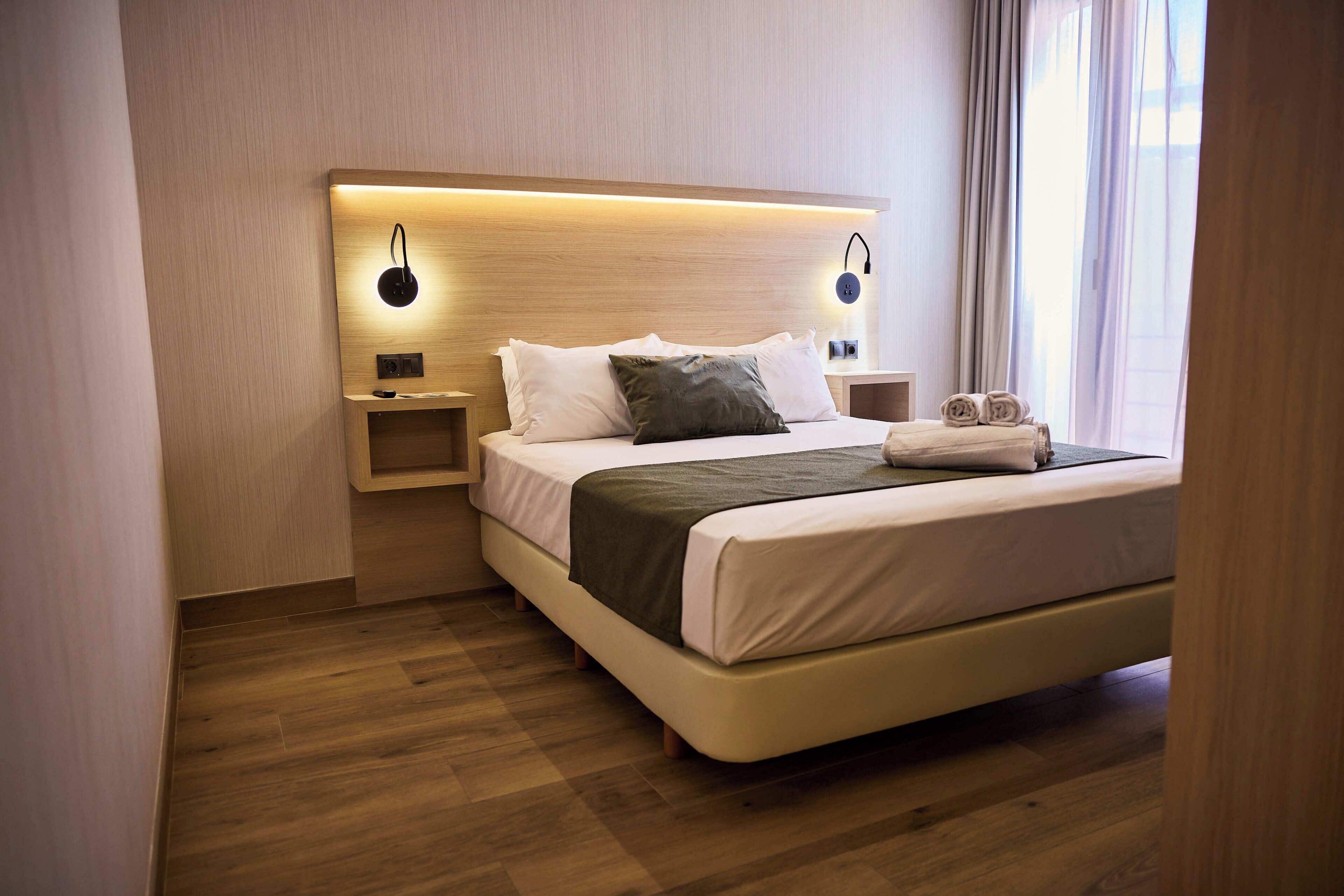 A modern hotel room with a large bed, bedside tables, and a headboard with integrated lighting.