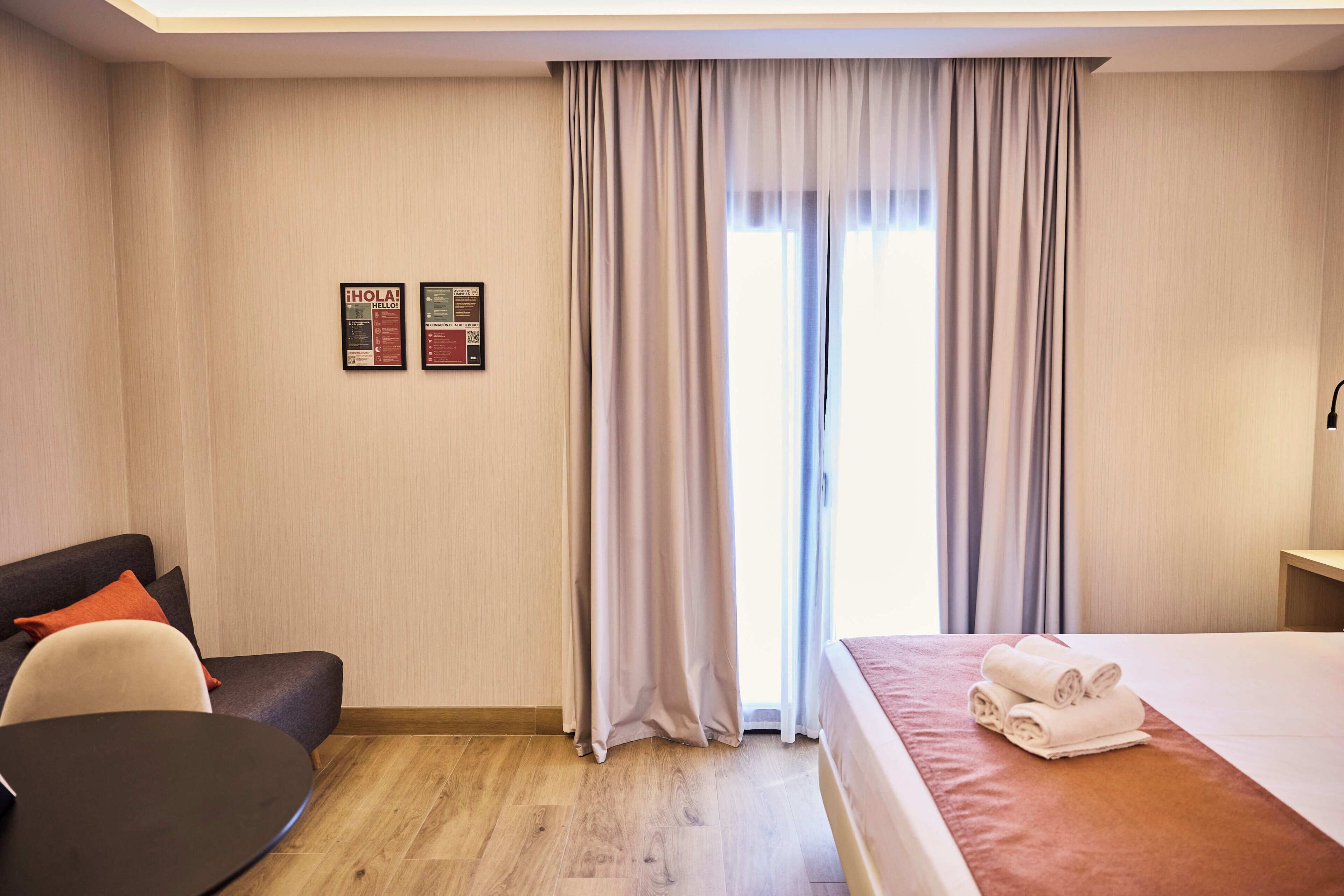 Ett hotellrum med en säng, ett skrivbord, en stol och ett litet bord. Det finns gardiner vid fönstret och två informationsaffischer på väggen.