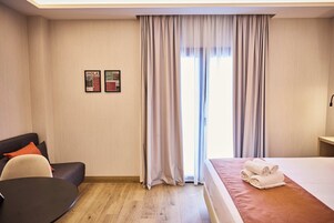Ein Hotelzimmer mit einem Bett, einem Schreibtisch, einem Stuhl und einem kleinen Tisch. Vor dem Fenster hängen Vorhänge und an der Wand befinden sich zwei Informationsplakate.