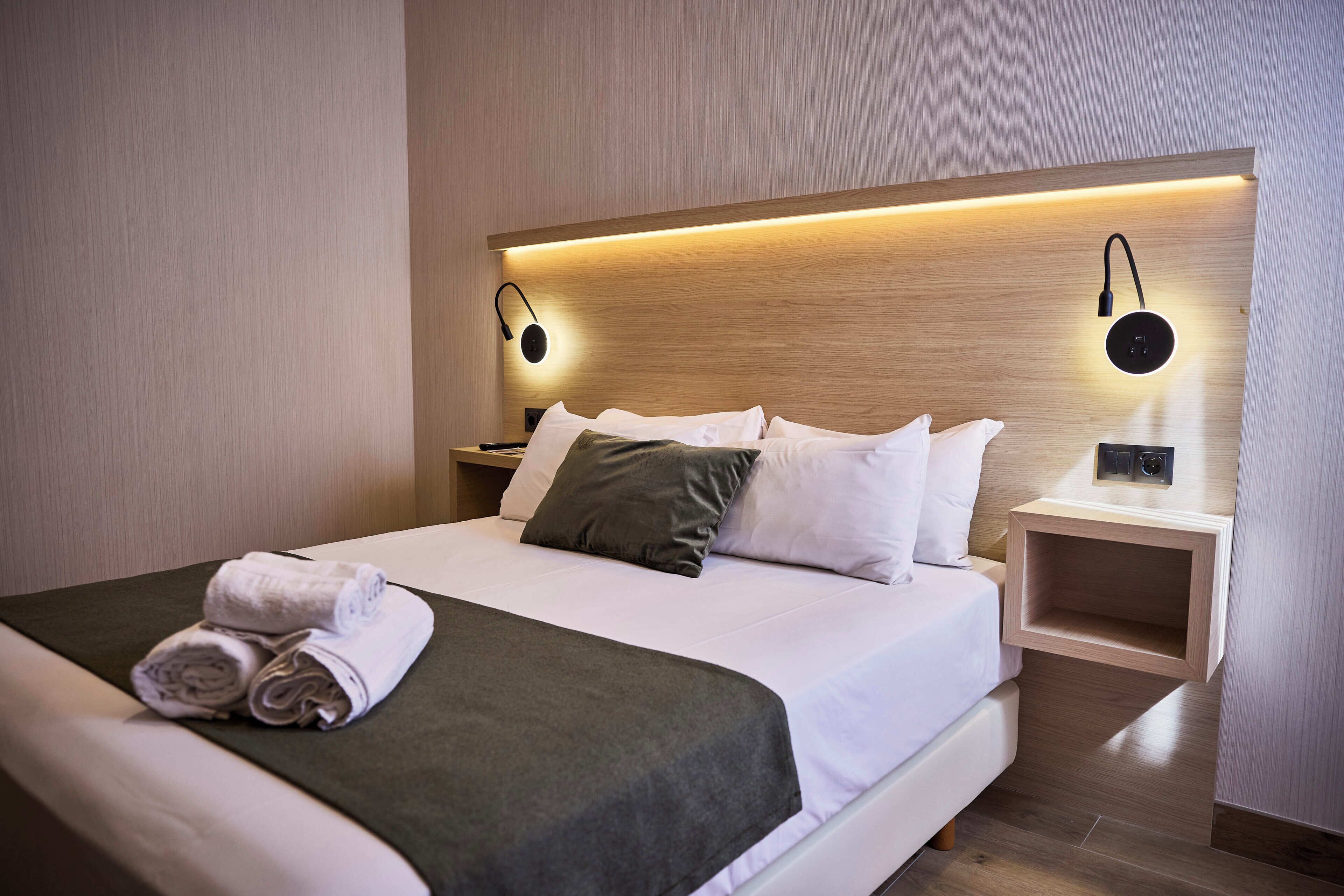A hotel room with a neatly made bed, two bedside tables, and a wall-mounted light fixture.