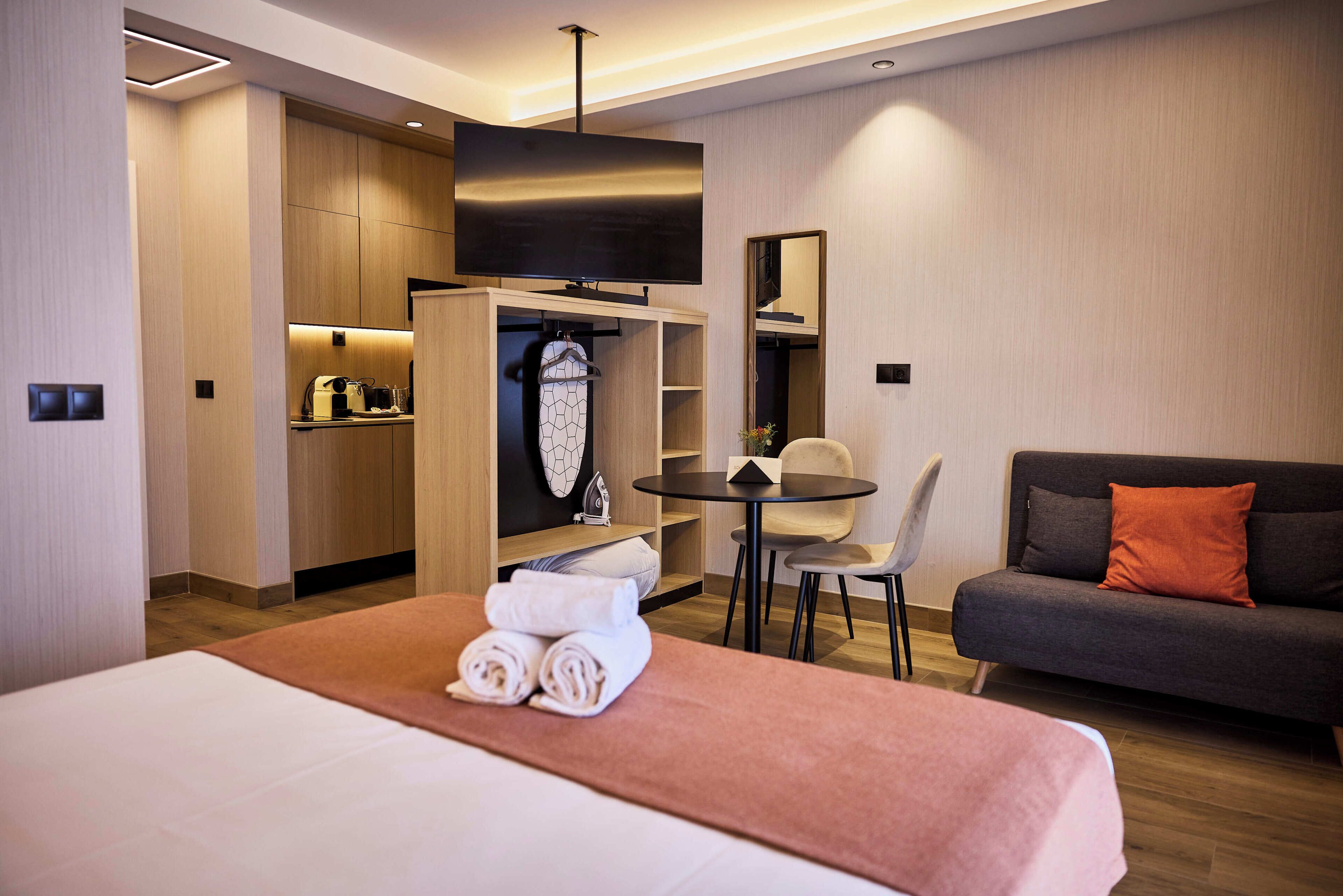 A hotel room with a bed, a sofa, a dining table with chairs, a TV, and a kitchenette.