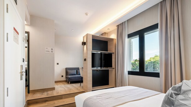 A modern hotel room with a bed, a seating area with a chair, a built-in TV, and a window with curtains.