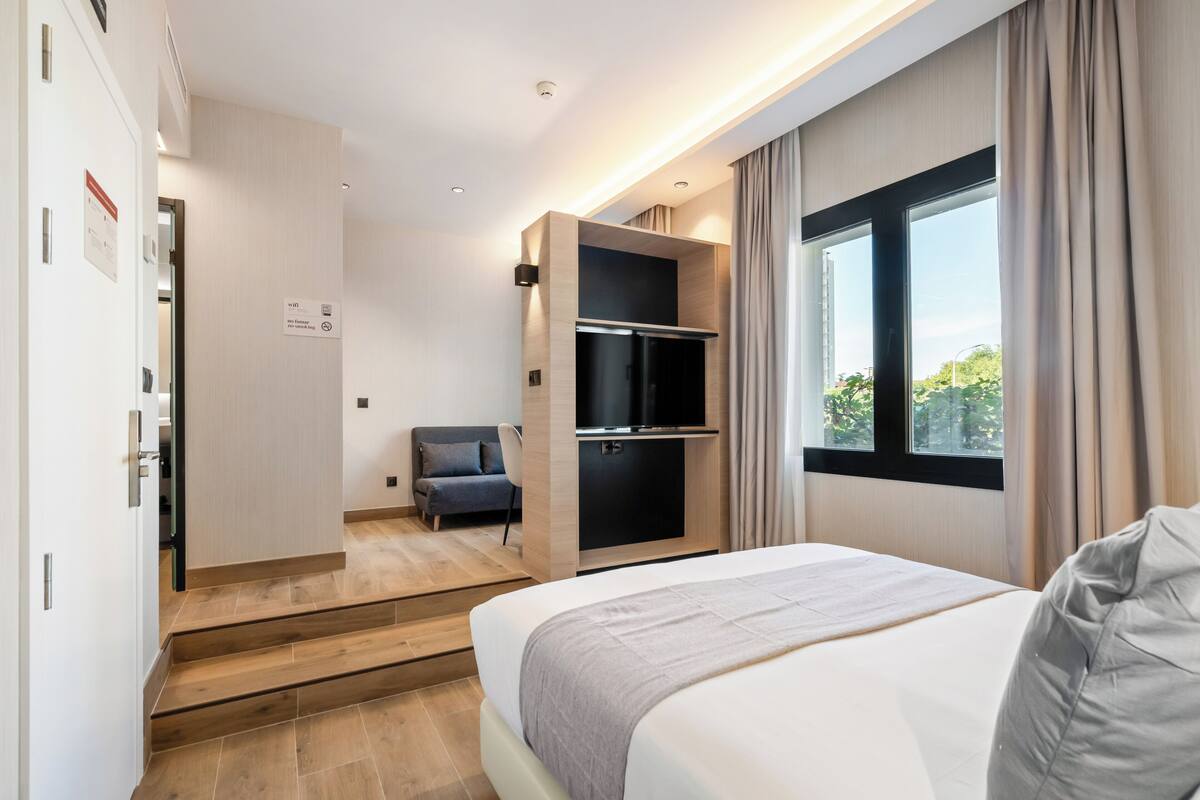A modern hotel room with a bed, a seating area with a chair, a built-in TV, and a window with curtains.