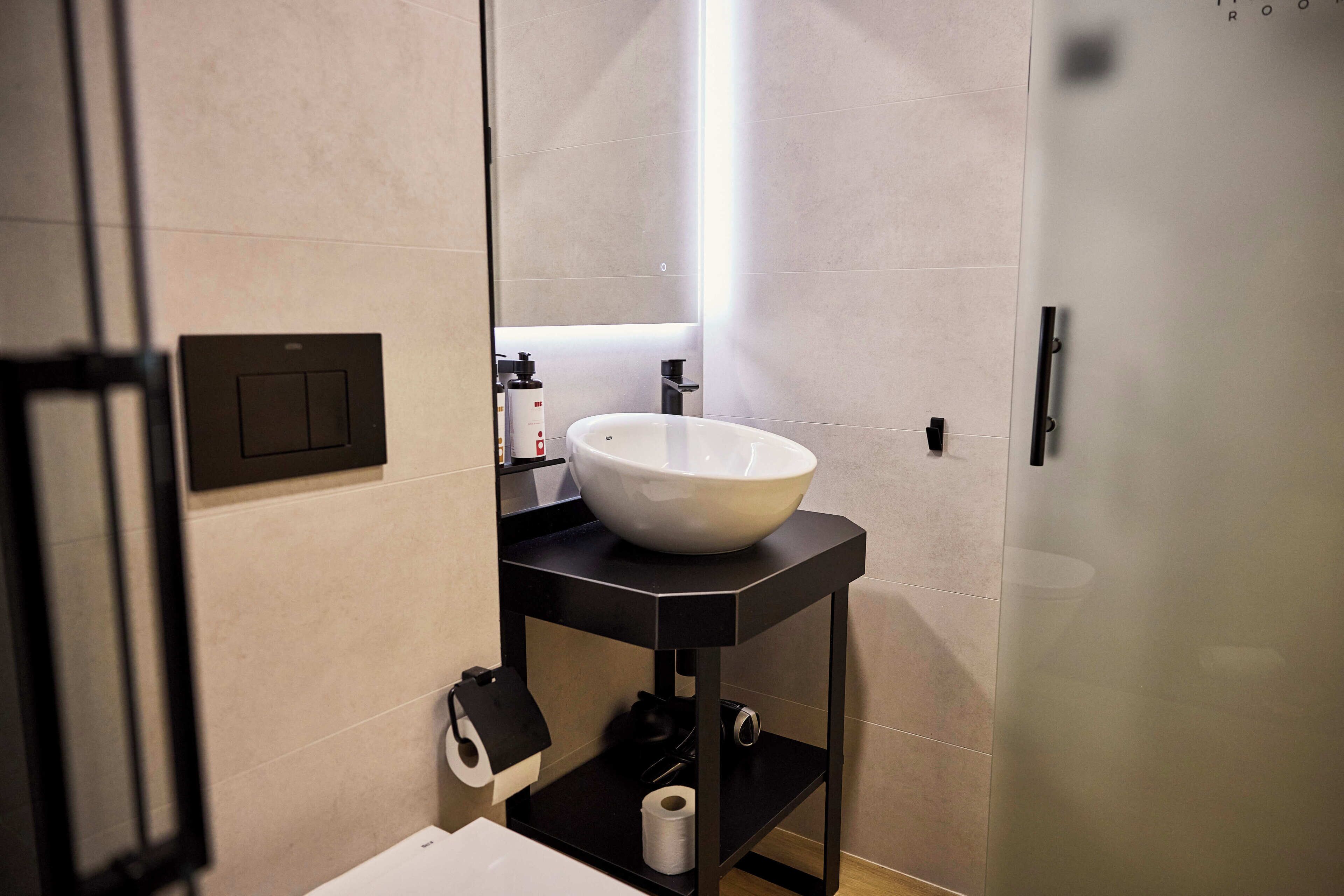 Un baño moderno con un lavamanos blanco sobre una encimera negra, un dispensador de papel higiénico y un perchero para toallas.