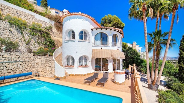 Una casa de dos pisos con piscina, tumbonas y un muro de piedra.