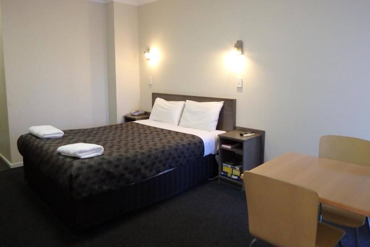 A hotel room with a bed, bedside table, chair, and wall-mounted lights.
