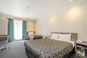 Quarto de hotel com uma cama grande, uma cama menor, uma cadeira, uma escrivaninha com telefone e uma estante de livros.
