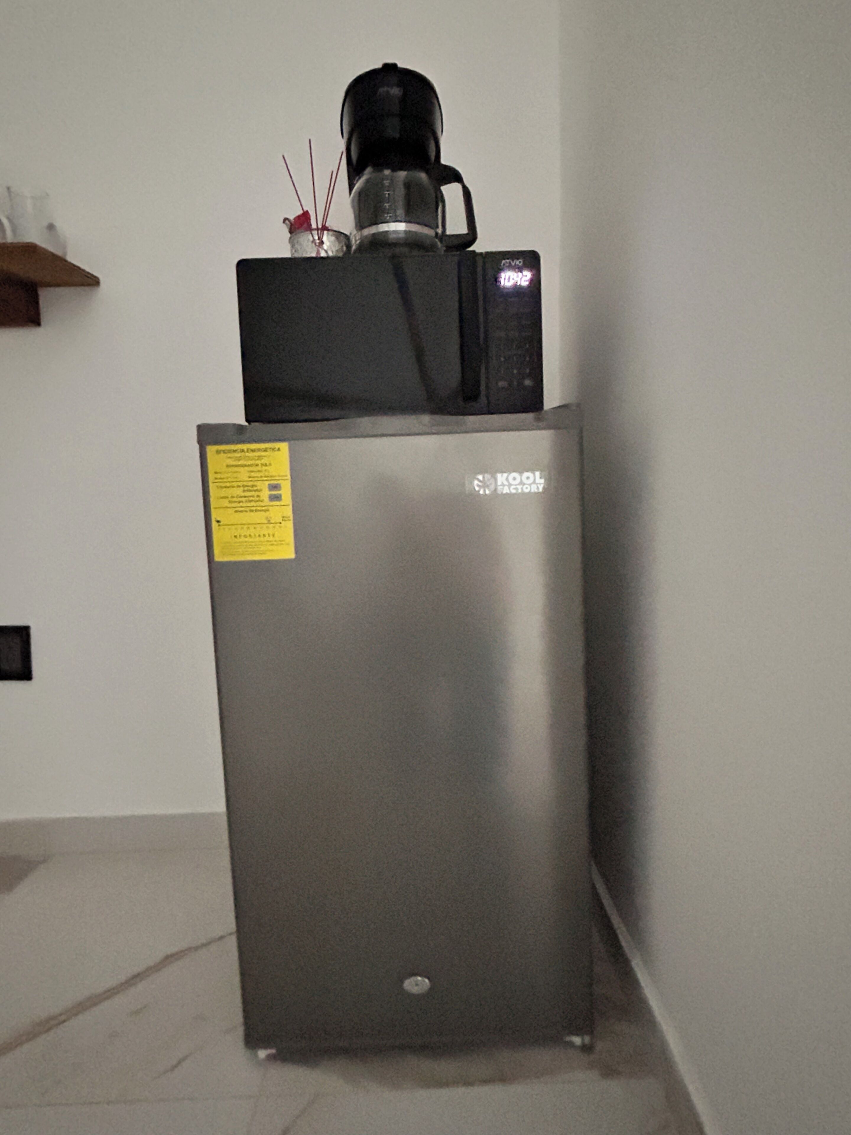 Un refrigerador de acero inoxidable con una cafetera encima, una etiqueta amarilla a la izquierda y un estante pequeño arriba.