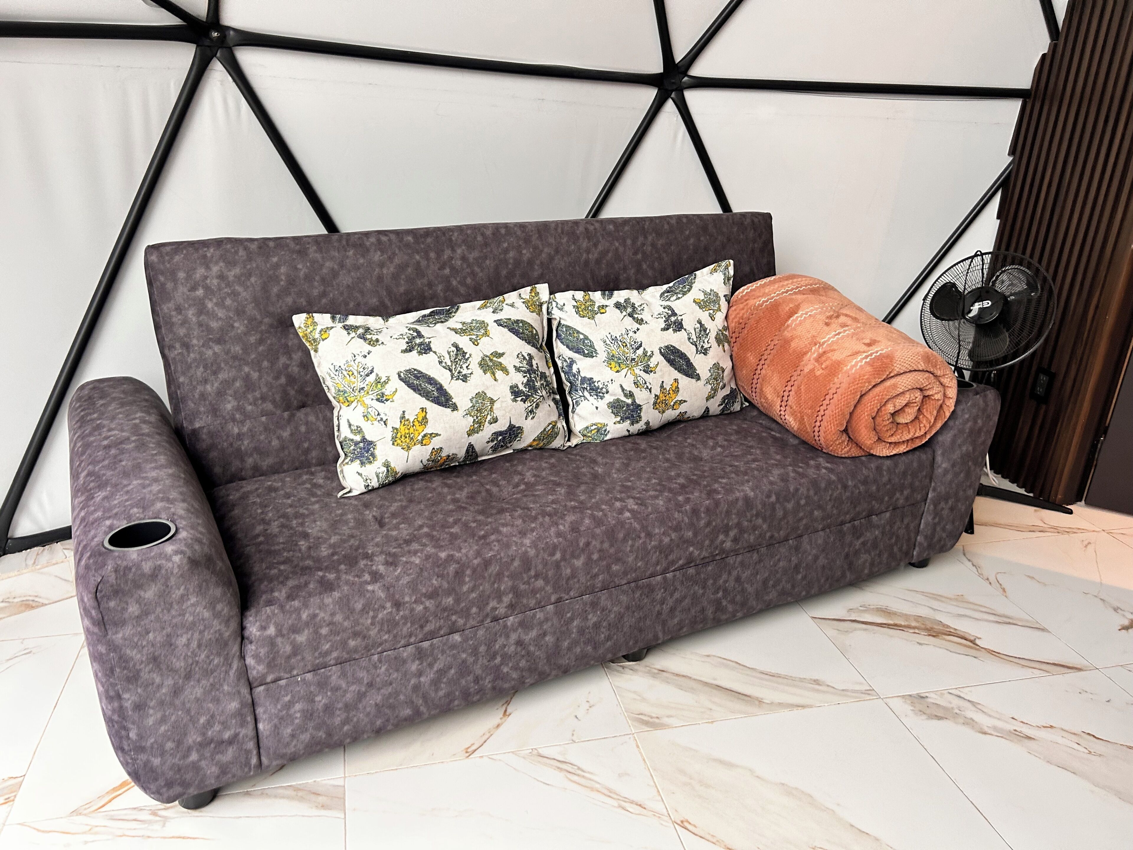 A grey sofa with patterned cushions and a rolled blanket, positioned against a wall with a geometric design.