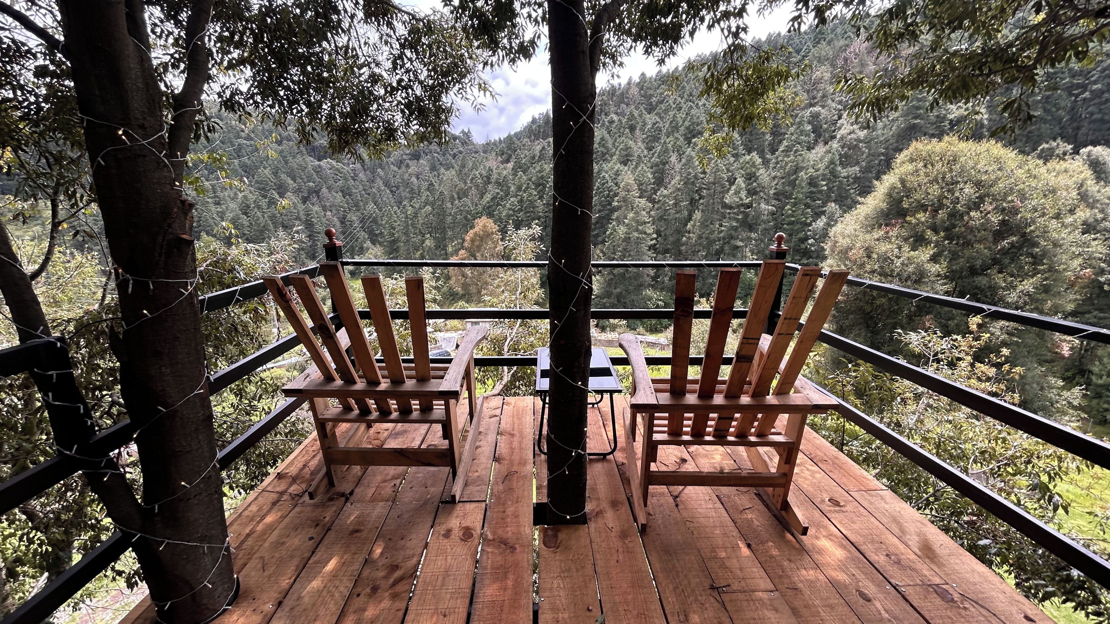 A wooden deck with two chairs and a table, surrounded by a metal railing, overlooking a forested hillside.