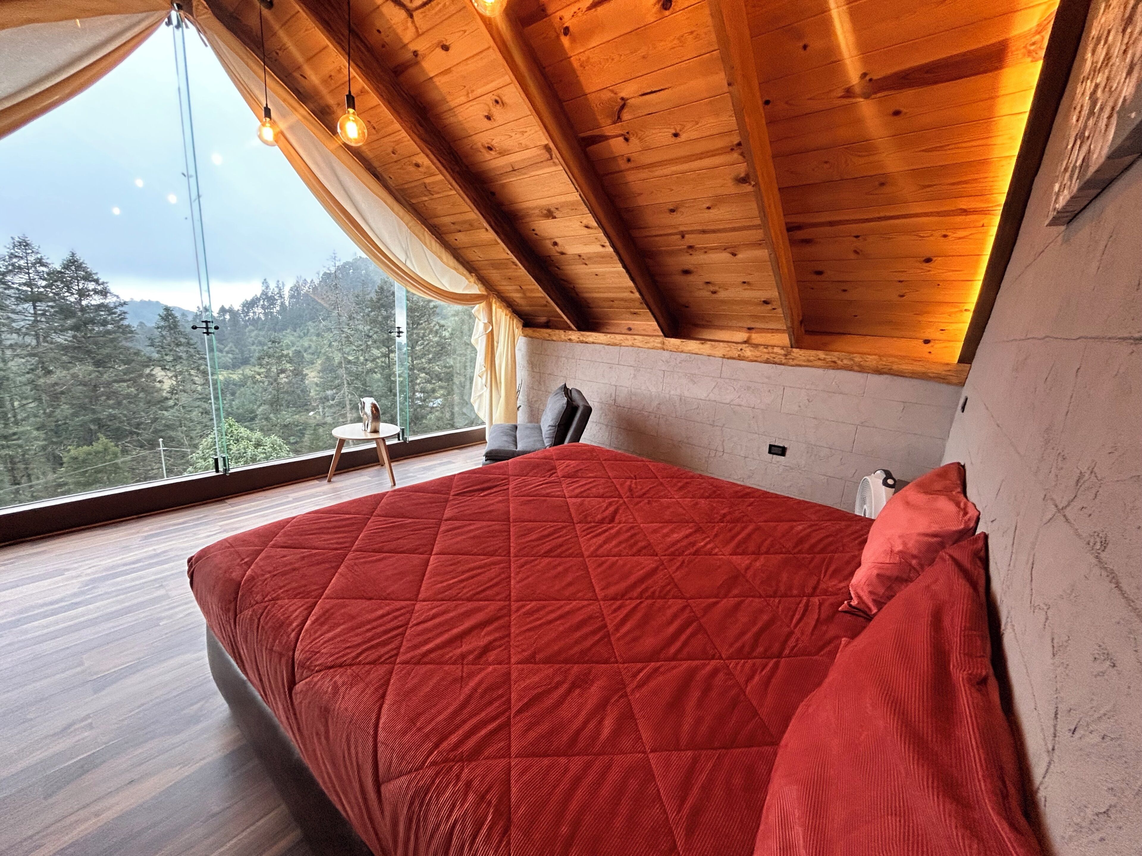 A bedroom with a wooden ceiling, a red bedspread, and a view of a forest through a large window.