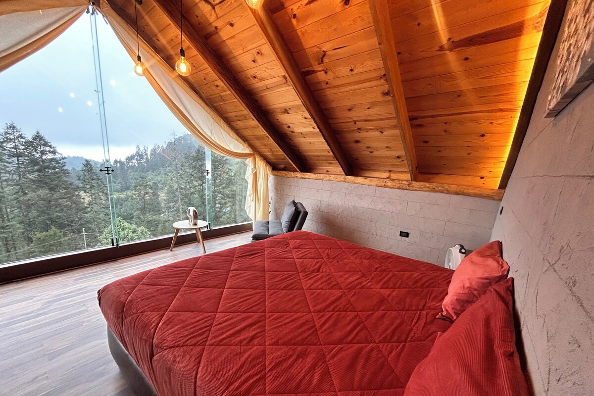 Un dormitorio con techo de madera, colcha roja y vista a un bosque a través de un ventanal grande.