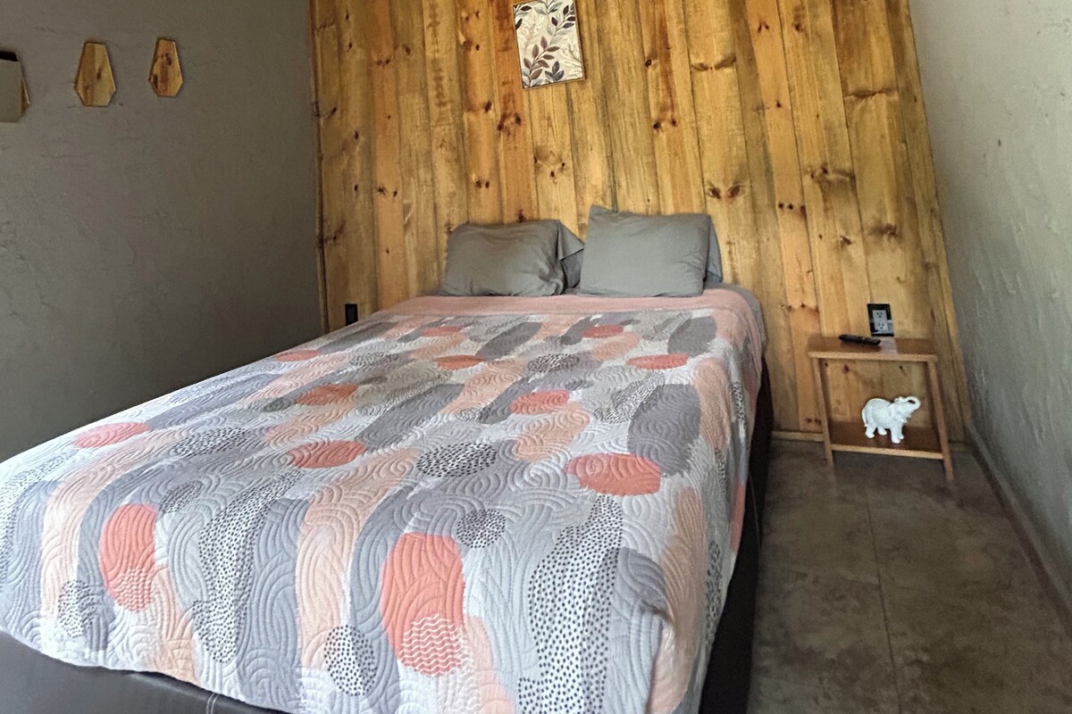 Un dormitorio con techo de madera, una cama con colcha estampada, dos almohadas, una mesita de noche con una figurita y un cuadro colgado en la pared.