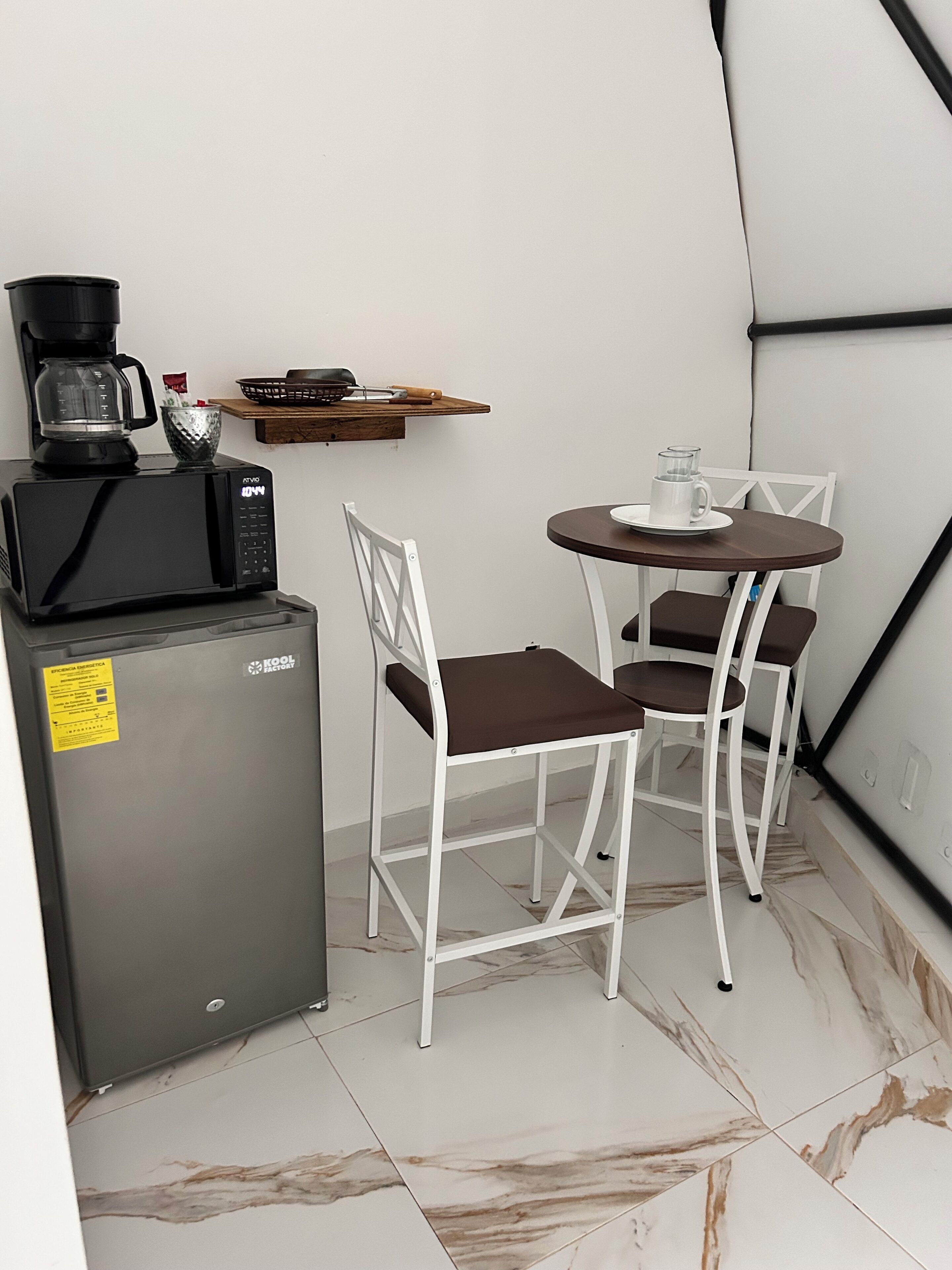 Une petite cuisine équipée d’un four à micro-ondes, d’une machine à café et d’un petit réfrigérateur. On y trouve une chaise blanche et une petite table ronde avec une tasse blanche.