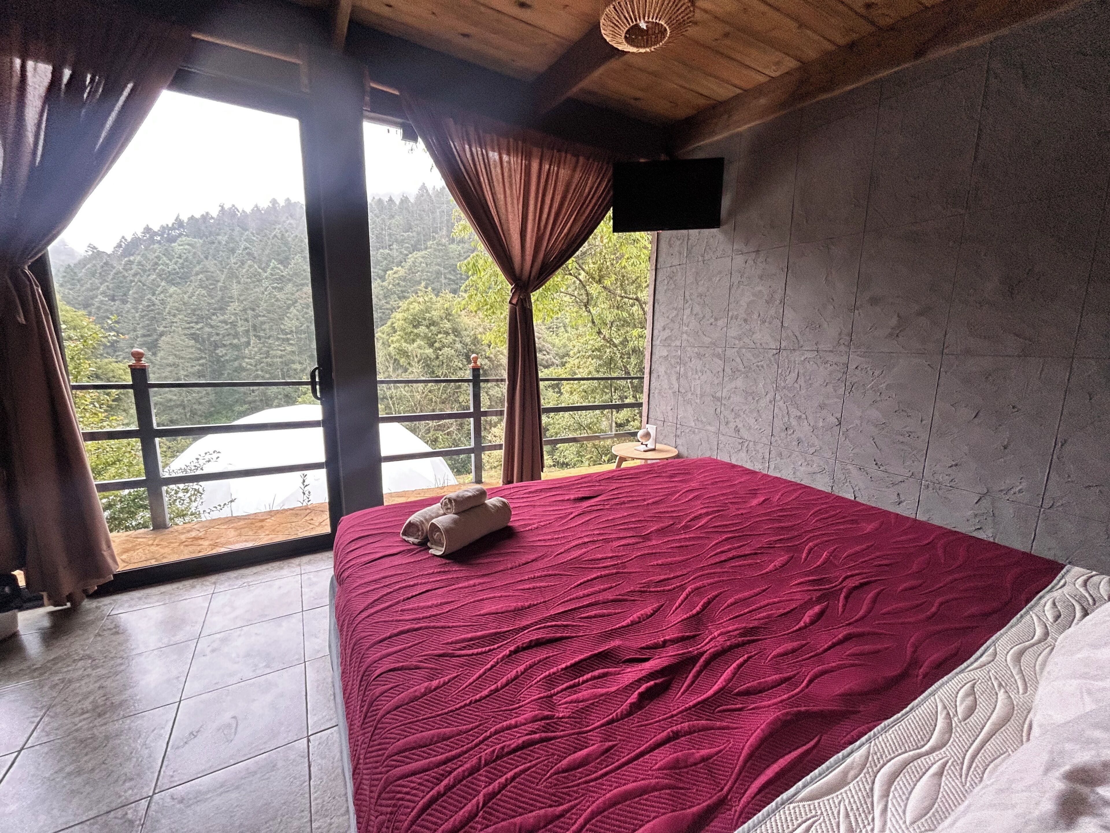 Quarto com cama grande, televisão na parede e varanda vista para a floresta.