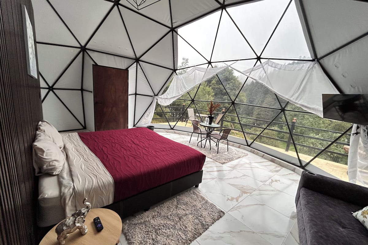 A bedroom with a bed, a small table, and a view of the outdoors through a geometric window structure.