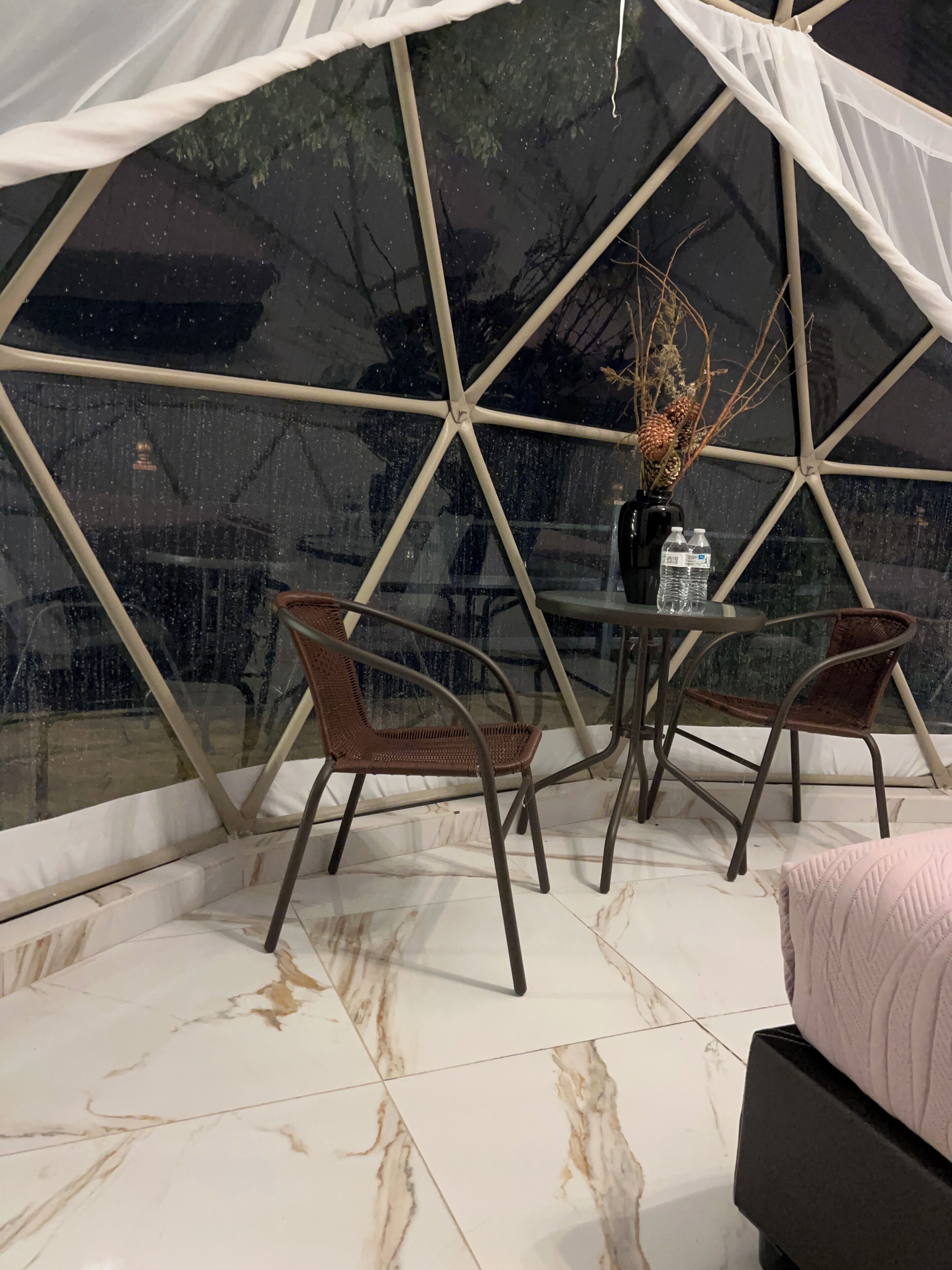 Una estructura tipo tienda con un ventanal de vidrio, una mesa redonda con sillas, un jarrón con plantas secas y una cama con una colcha rosa.
