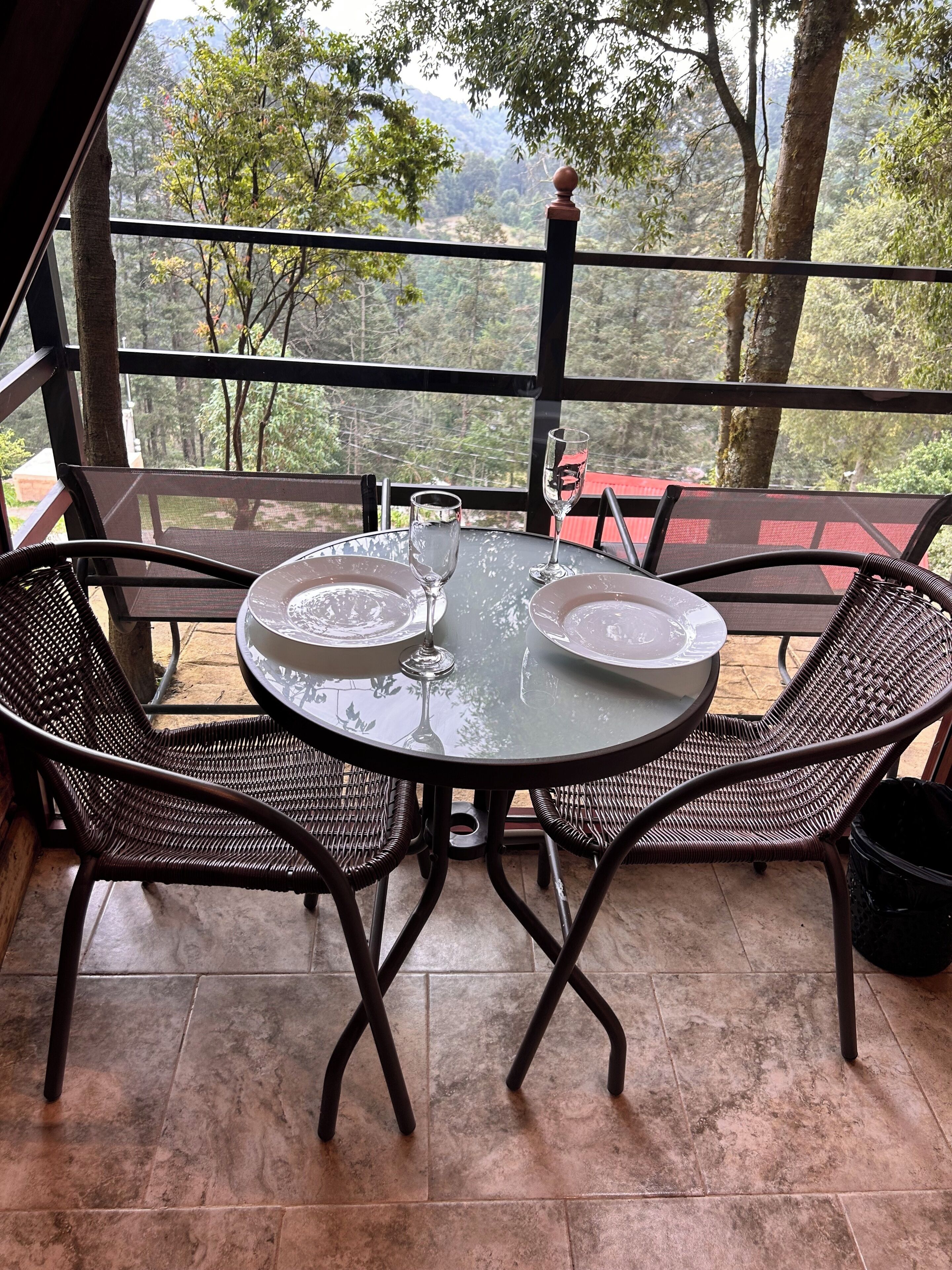 Mesa de vidrio redonda con dos sillas, un vaso y un plato en un balcón con vista a árboles y montañas.