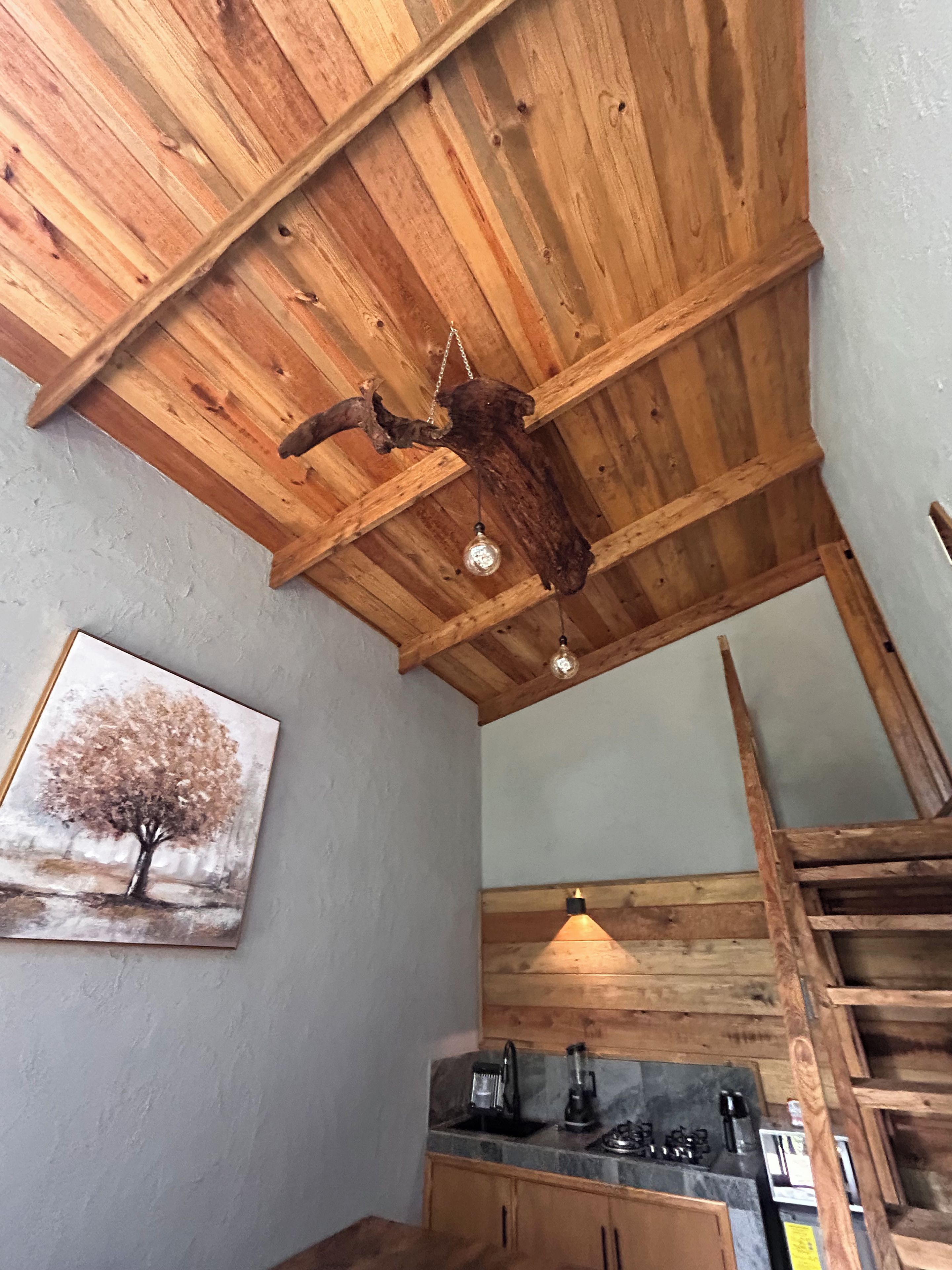 A rustic kitchen with wooden beams, a hanging light fixture, and a painting of a tree.