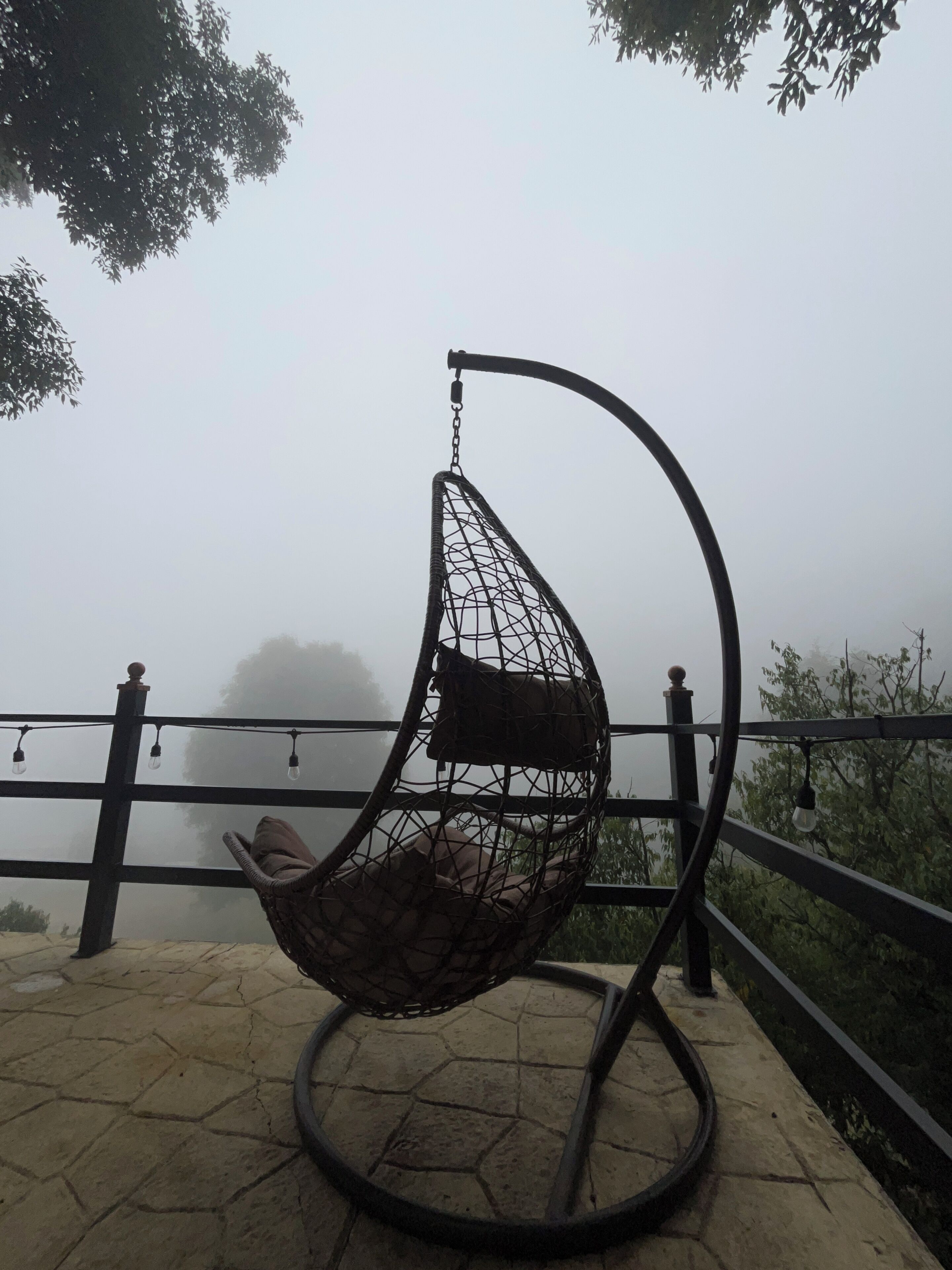 Una poltrona sospesa con seduta e schienale a rete, posizionata su un patio in pietra con ringhiera e luci decorative.