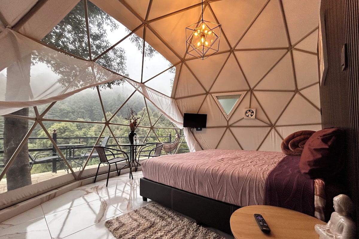 A room with a bed, a small table, a chair, a TV, and a view of the outdoors.