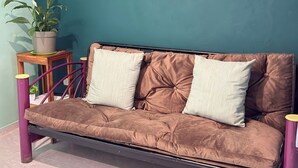 A brown sofa with two white cushions against a teal wall, a small wooden table with a potted plant, and purple cylindrical stools.
