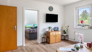 A room with a wooden door, a television mounted on the wall, a small cabinet with drawers, a window with a view of greenery, and a bed with striped pillows.