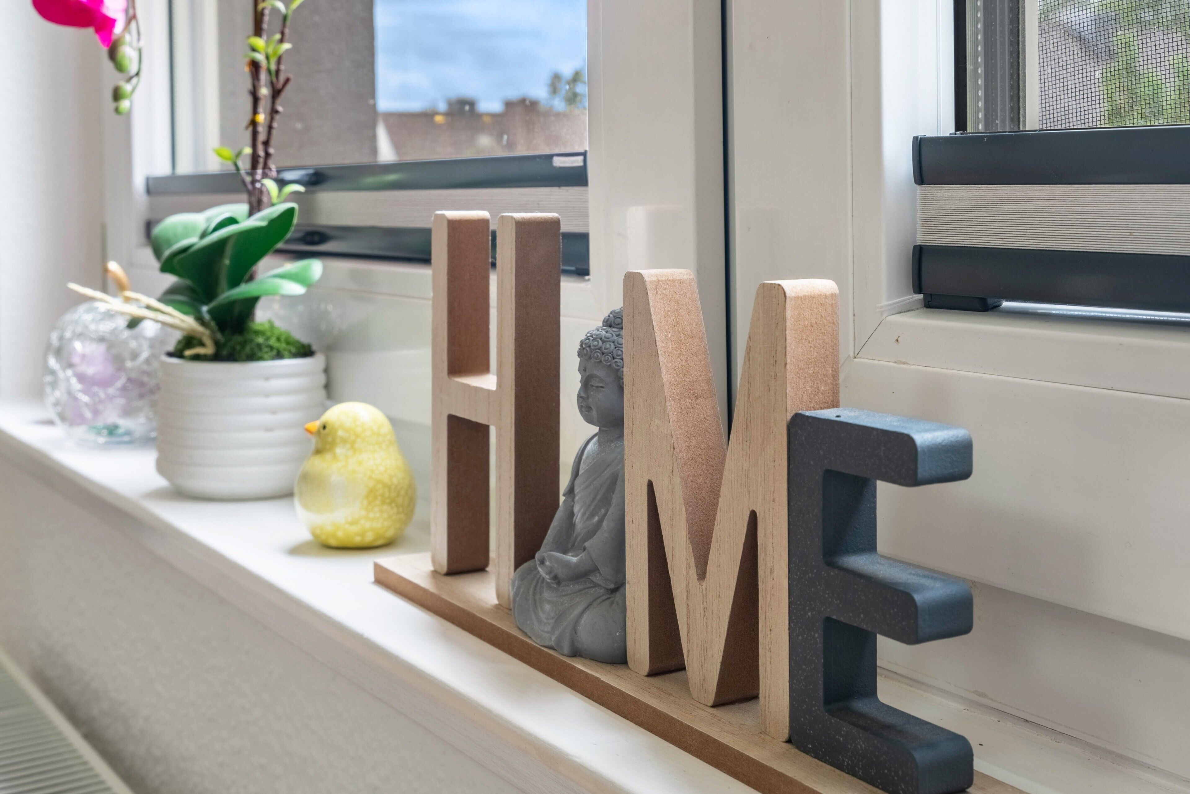 Eine Fensterbank mit Dekorationsobjekten, darunter Holzbuchstaben, die ‚HOME‘ buchstabieren, und eine kleine Buddha-Statue.