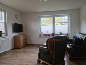 Un salon avec un meuble en bois, un téléviseur, un fauteuil vert et une fenêtre donnant sur un paysage verdoyant.