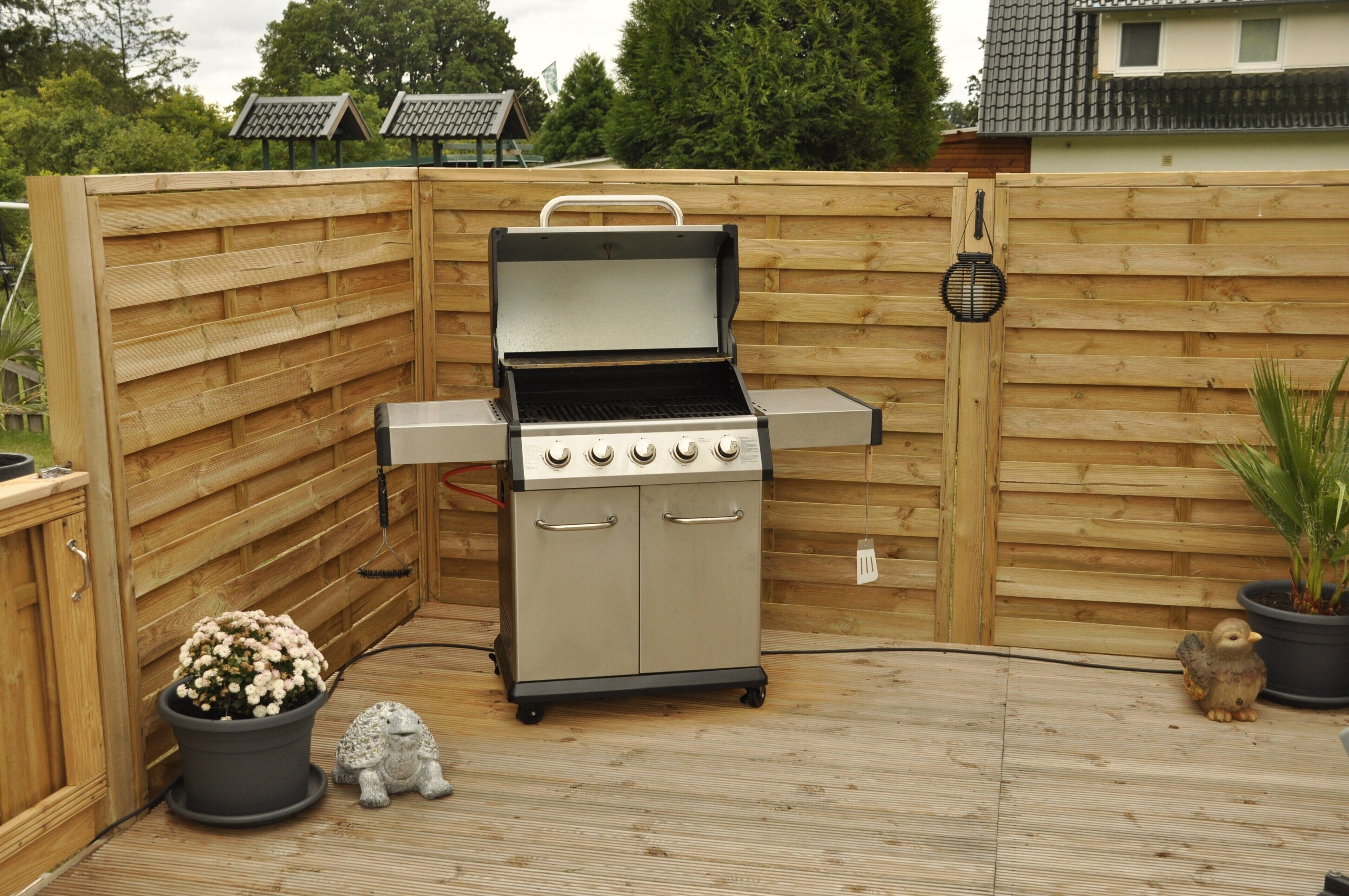 Una parrilla de gas portátil con cuatro quemadores, un quemador lateral y un cajón de almacenamiento, montada en un patio trasero con cerco de madera.