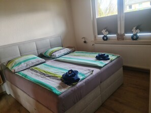 Ein Doppelzimmer mit zwei Betten, einem Fenster mit Jalousien, einem Heizkörper und einer kleinen Ablagefläche mit Dekoration.