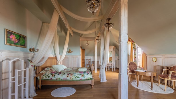 A spacious bedroom with a large bed, a wooden floor, a chandelier, and a view of the hallway. - Mazmežotnes muiža (Mazmežotne)