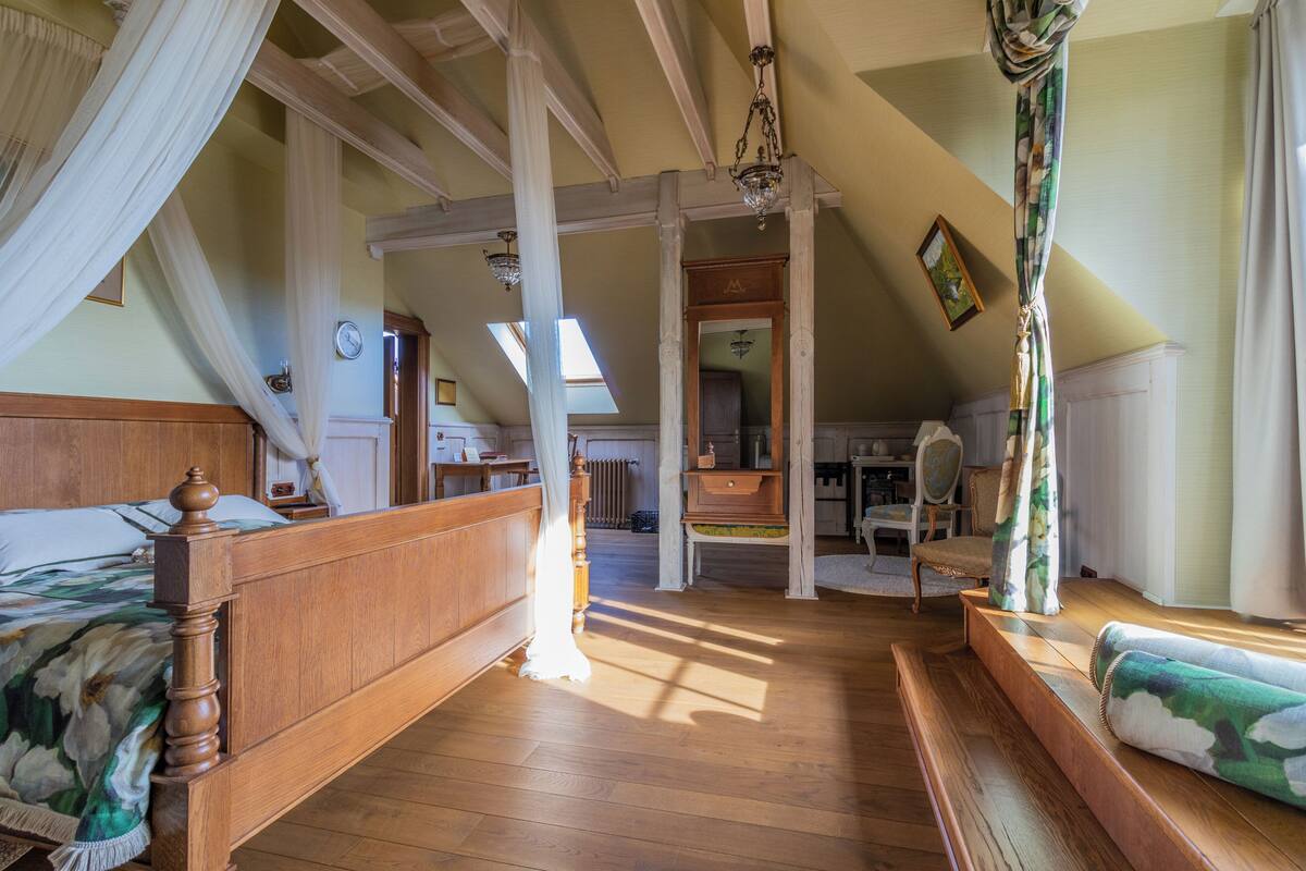 Un dormitorio con una cama de madera, un banco, una silla y vista a la cocina.