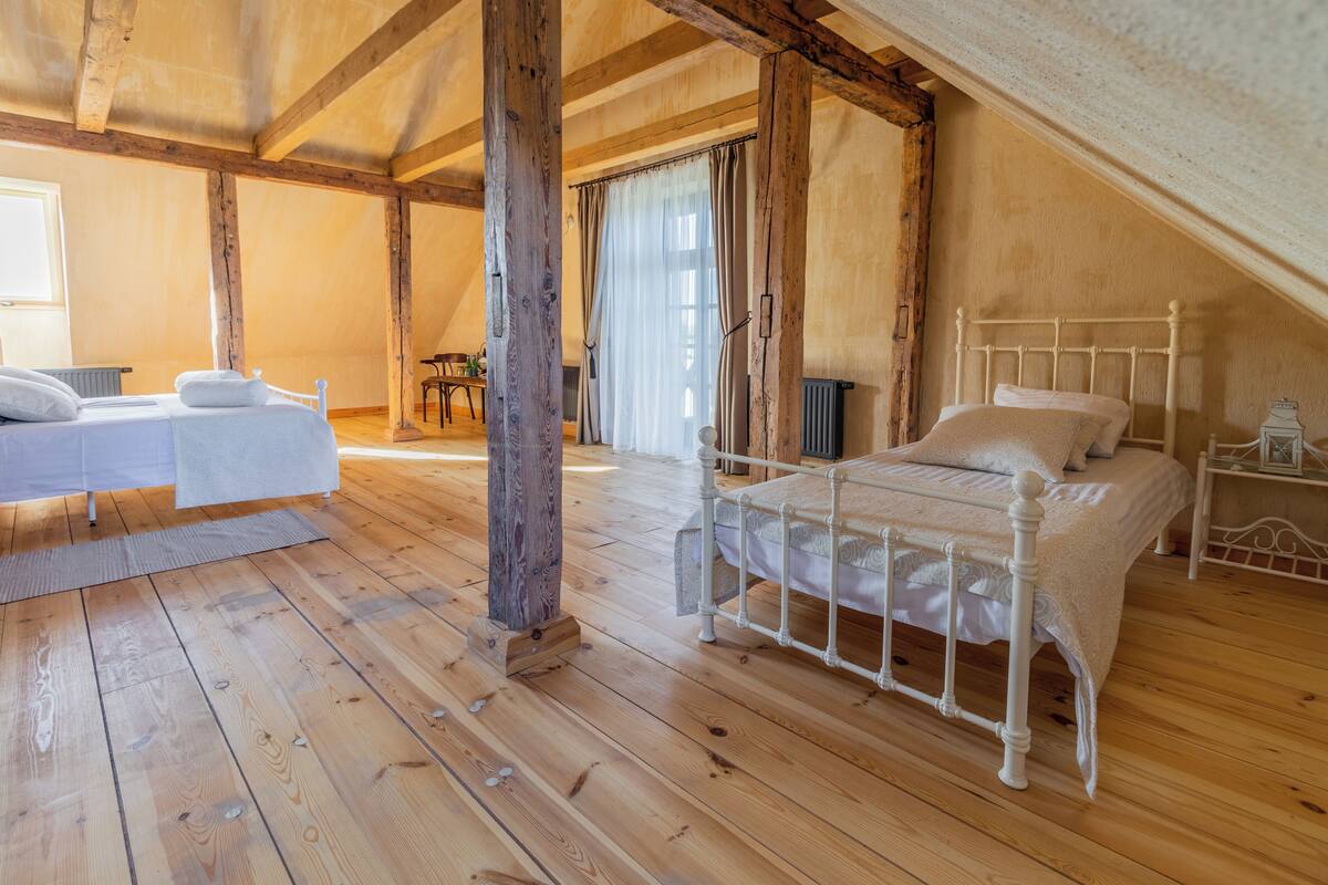 Una habitación con techo inclinado, vigas de madera, una cama con cabecero blanco, una mesita de noche y una ventana con cortinas.