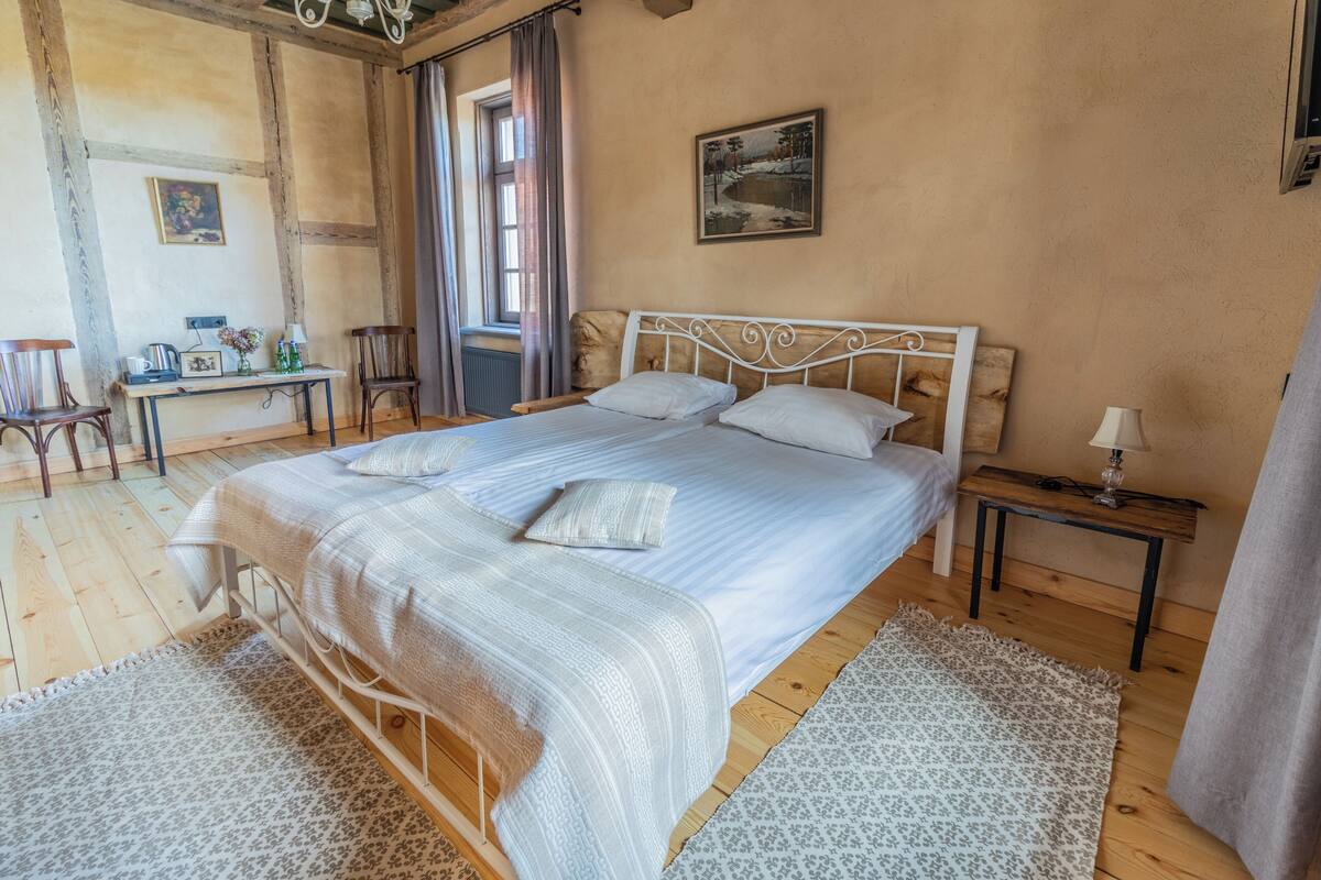 Un dormitorio con una cama grande, suelo de madera, una mesita con lámpara y un cuadro en la pared.